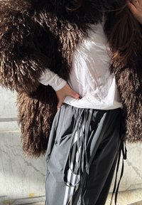 Personne portant un manteau brun à poils longs, une chemise blanche à manches longues et un pantalon gris avec des rubans rayés noirs et blancs, main posée sur la hanche.