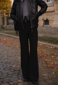 Czarna skórzana kurtka bomberka na szarym swetrze, czarne, rozszerzane spodnie z wysokim stanem, błyszczące buty, stojąca na bruku z jesiennymi liśćmi.