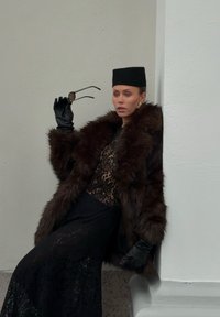 Brun pelskåpe i kunstpels over en svart blonderkjole. Modellen holder runde solbriller, iført svarte skinnhansker, foran en lys grå vegg.