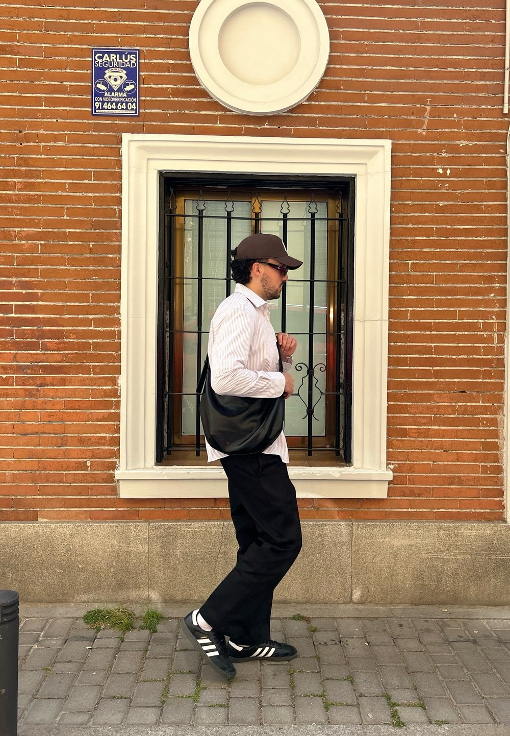 Borsa nera a tracolla, camicia bianca a righe, pantaloni scuri e scarpe nere con strisce bianche. Muro in mattoni rossi con una finestra e barre nere.