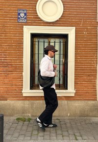 Borsa nera a tracolla, camicia bianca a righe, pantaloni scuri e scarpe nere con strisce bianche. Muro in mattoni rossi con una finestra e barre nere.