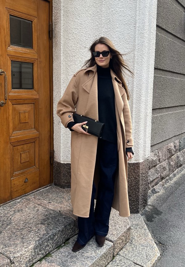 Abrigo de lana beige sobre un suéter negro de cuello alto, combinado con pantalones de mezclilla oscuros de campana. Sosteniendo un bolso de mano negro con acentos dorados. Zapatos marrones de punta.