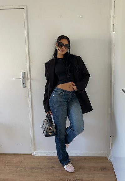 Mujer con top corto negro, blazer negro de gran tamaño, vaqueros azules, zapatos blancos, gafas de sol grandes, sosteniendo un bolso negro en el interior.