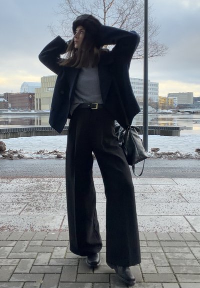 Mujer con pantalones negros de pierna ancha, abrigo negro y sombrero de piel posando en una acera de la ciudad, con edificios nevados y agua al fondo.