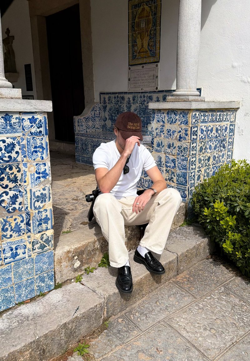 Mann in weißem Hemd und beiger Hose sitzt auf Steinstufen neben einer blau-weißen, gefliesten Wand, richtet seinen braunen Hut und hat eine Kamera neben sich liegen.