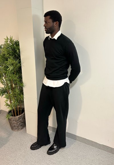 Hombre con barba y peinado afro de pie en el interior, vistiendo suéter negro, camisa blanca, pantalones y zapatos negros junto a una planta verde en maceta.