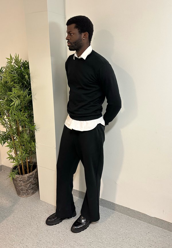 Man with beard and afro hairstyle stands indoors wearing black jumper, white shirt, black trousers and shoes beside a potted green plant.