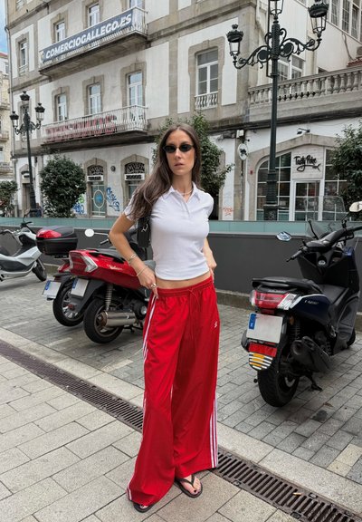 Pantalones deportivos anchos rojos con rayas laterales blancas, combinados con una camiseta polo ajustada blanca y gafas de sol negras. Al fondo, hay scooters aparcados.