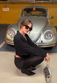 Črn skrajšan blazer, prugasta mini krila, prozorne črne nogavice, konični črni škornji, prozorna torba s kovinskimi detajli, srebrni vintage avtomobil v ozadju.