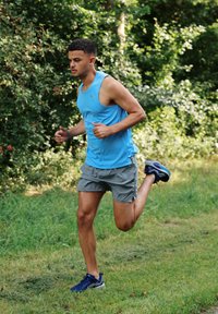 En atletisk mand løber i en lyseblå tanktop og grå shorts, iført blå løbesko, med en kort frisure, omgivet af grønne omgivelser.