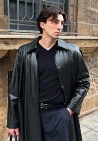 Homme portant un manteau en cuir noir texturé, un pull noir et un pantalon à rayures fines, debout près d'un mur en pierre avec une fenêtre à barreaux en métal.