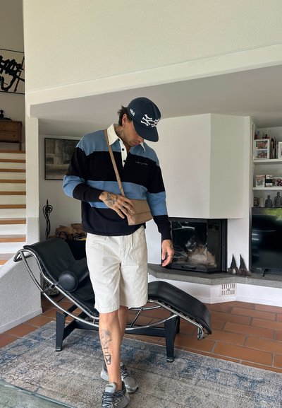 Suéter a rayas en negro, azul y blanco, con una bolsa de hombro beige, pantalones cortos claros, zapatillas grises y una gorra azul marino, de pie en el interior.