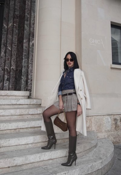 Abrigo crema, camisa de mezclilla, falda de cuadros, bolso marrón, medias oscuras y botas altas verdes con tacones. Escaleras de mármol y fondo de pared beige.