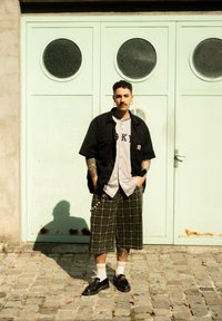 Fekete rövid ujjú póló egy szürke baseball mez felett, melyen "OK" felirat látható, zöld kockás rövidnadrág, fekete cipő és fehér zokni.