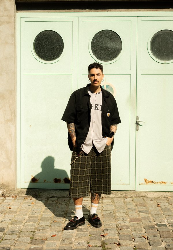 Chemise noire à manches courtes sur un maillot de baseball gris avec l'inscription "OK", shorts à carreaux verts, chaussures noires et chaussettes blanches.