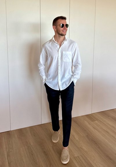 Joven con gafas de sol, camisa blanca abotonada, pantalones negros y zapatos beige, de pie en el interior sobre suelo de madera contra una pared blanca.