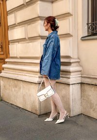 Džínová bunda, biele čipkované pančuchy a biele sandále na opätku. Drží bielu kabelku so zlatým akcentom. Hladké, štruktúrované materiály.