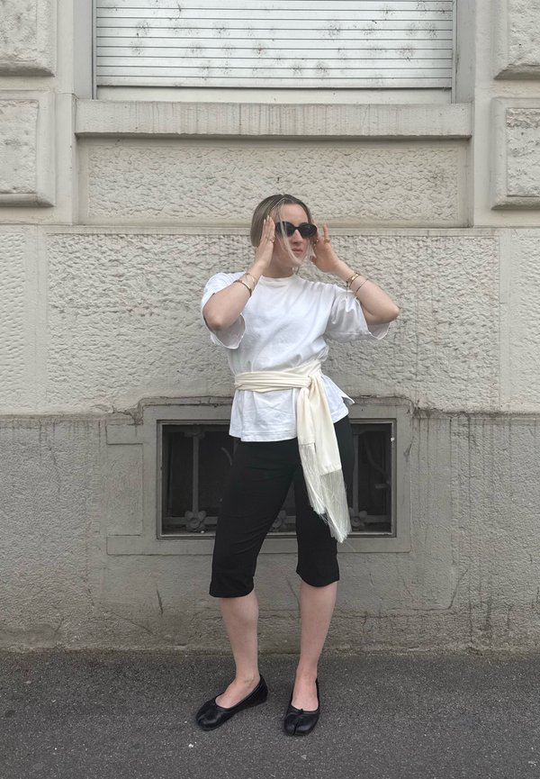 Persona con top bianco e pantaloni capri neri regola gli occhiali da sole mentre è in piedi per strada davanti a un muro di pietra con texture.