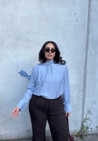 Blusa de mangas longas azul clara com decote franzido, combinada com calças largas castanhas escuras, complementada por óculos de sol pretos pequenos.