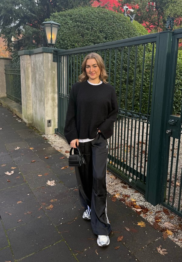 Joven mujer con suéter negro y pantalones deportivos Adidas, de pie en la acera junto a una puerta de hierro verde, sosteniendo un bolso pequeño negro, sonriendo.