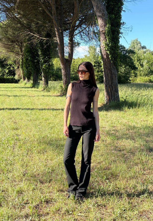 Femme portant un haut sans manches noir et un pantalon, debout dans une zone herbeuse ensoleillée avec de grands arbres en arrière-plan, portant de grandes lunettes de soleil.
