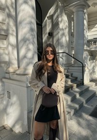 Μπέζ παλτό τέντσα πάνω από μαύρο φόρεμα, συνδυασμένο με σκούρα γυαλιά ηλίου. Ένα μικρό καφέ τσαντάκι κρατιέται μπροστά. Στο φόντο διακρίνονται λευκές αρχιτεκτονικές κατασκευές.