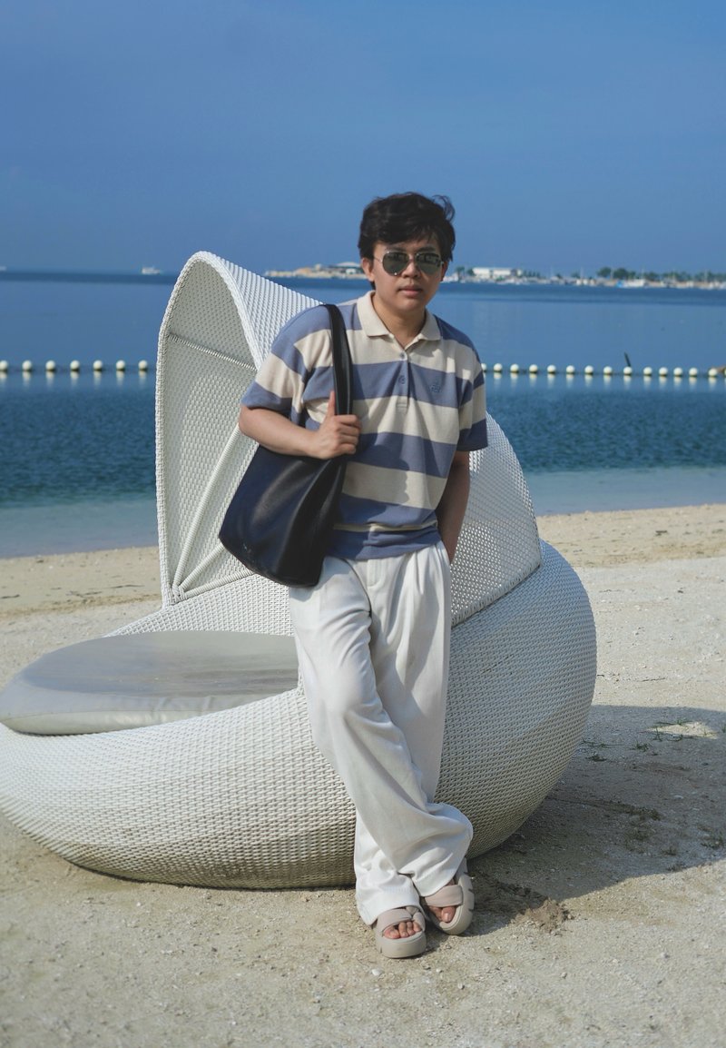Personne portant des lunettes de soleil et une chemise rayée se tient sur une plage de sable à côté d'une chaise longue blanche en osier près d'une eau calme sous un ciel dégagé.