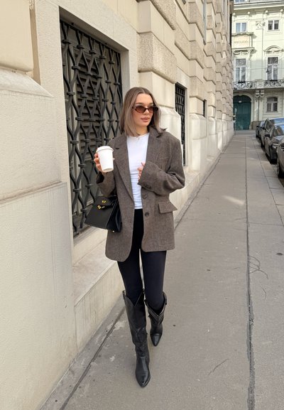 Blazer marrón oversized sobre una blusa blanca, combinado con leggings negros y botas de tacón puntiagudas hasta la rodilla. Bolso negro y taza de café en la mano.