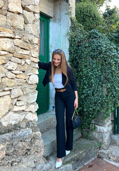 Blazer negro y pantalones de campana negros sobre una blusa blanca. Tacones blancos y un pequeño bolso negro. Muro de piedra y puerta verde de fondo.