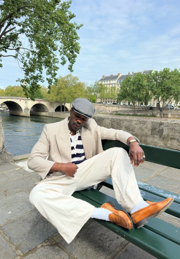 Homme en veste beige, pantalon blanc et chaussures orange assis détendu sur un banc vert près de la rivière avec un pont en pierre et des arbres en arrière-plan.