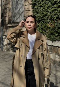 Manteau beige avec un grand col et un devant croisé, porté sur un t-shirt blanc et un pantalon noir, se tenant dans une rue ensoleillée.