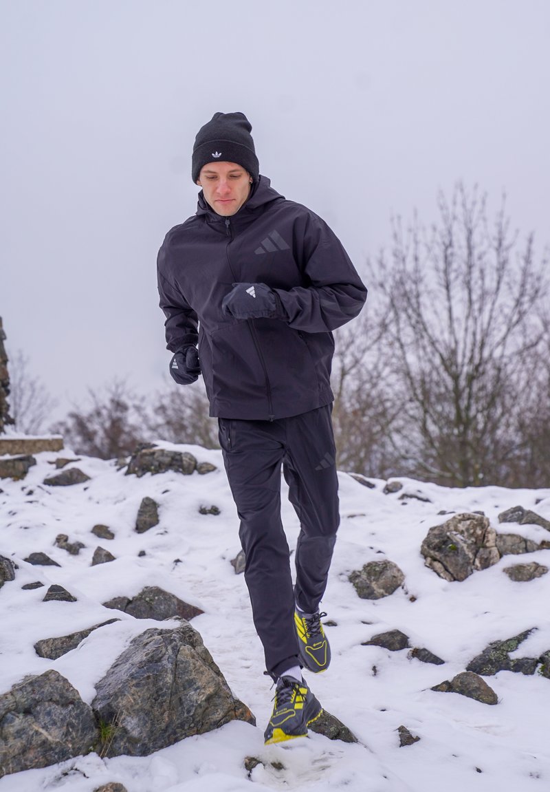 Homme vêtu d'un équipement d'hiver noir courant sur un terrain enneigé et rocheux, avec des arbres sans feuilles en arrière-plan sous un ciel gris.