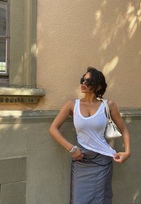 Žena v slnečných okuliaroch, bielom topu, sivých sukni a strieborných náramkoch stojí proti béžovej stene a jednou rukou si upravuje top.