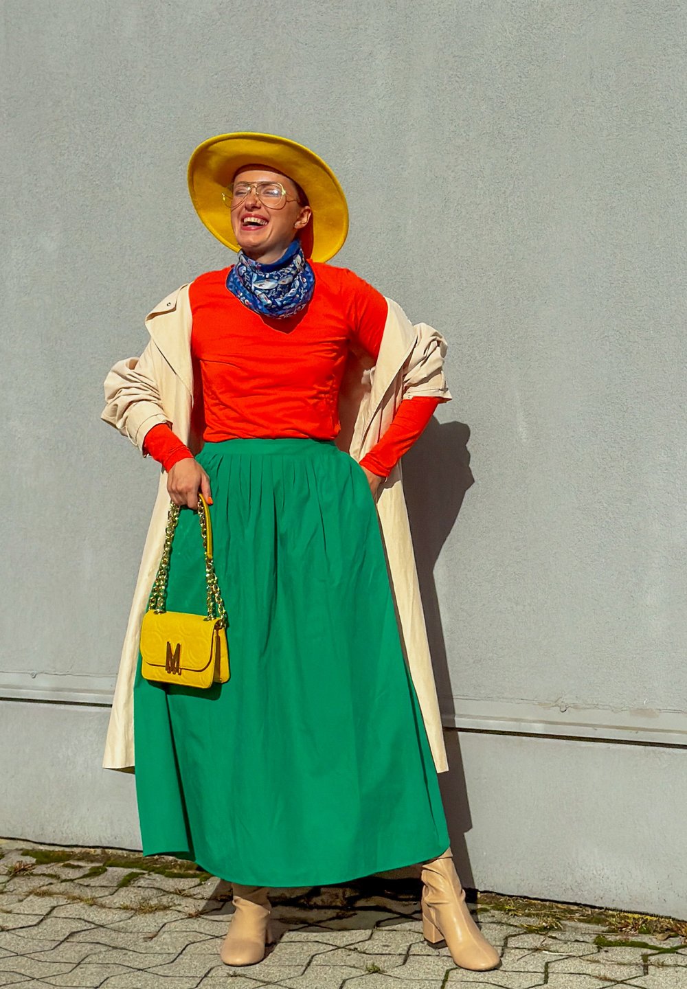 Helle gelbe breitbrimige Hut, orangefarbene langärmelige Bluse, grüne knielange Rock, hellbeige kniehohe Stiefel und eine kleine gelbe Handtasche.