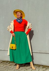 Helle gelbe breitbrimige Hut, orangefarbene langärmelige Bluse, grüne knielange Rock, hellbeige kniehohe Stiefel und eine kleine gelbe Handtasche.