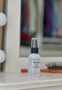 Σπρέι παχύτητας Bumble and bumble σε φ frosted γυάλινο μπουκάλι με μαύρη αντλία στην κορυφή, ετικετοποιημένο με λευκό κείμενο. Κοντά υπάρχει μια πορτοκαλί χτένα.