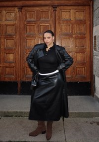 Abrigo largo de cuero negro sobre una camiseta negra, combinado con una falda de cuero negra. Botines marrones y un bolso pequeño completan el conjunto. Puertas de madera al fondo.