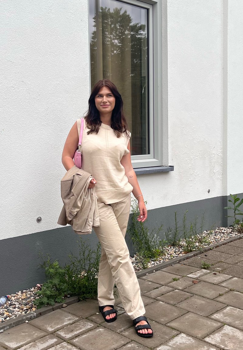 Frau mit dunklen Haaren läuft auf einem Steinweg, trägt einen beige ärmellosen Pullover und Hose, schwarze Sandalen, hält eine beige Jacke und eine pinke Tasche.