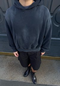 Άτομο που φοράει σκούρο φαρδύ φούτερ με κουκούλα, μαύρο σορτς και μαύρα μποτάκια μέχρι τον αστράγαλο, στέκεται σε υφή εδάφους μπροστά σε έναν σκοτεινό πορτάκι.