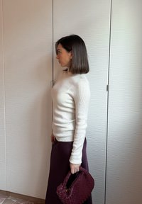 Femme aux cheveux bruns mi-longs portant un pull blanc et une jupe foncée, tenant un sac à main tissé rouge foncé, debout de profil à l'intérieur.