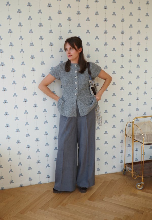 Mujer con suéter gris de manga corta abotonado y pantalones grises anchos, de pie con las manos en las caderas frente a un papel pintado floral.