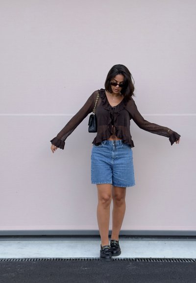 Top largo de manga larga marrón transparente con volantes, combinado con shorts de mezclilla azul claro. Un bolso de hombro negro y zapatillas negras completan el conjunto.