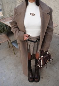 Manteau en laine marron, col roulé côtelé blanc, jupe plissée à carreaux, bottes marron montant jusqu'aux genoux, et sac à main marron avec des accents.