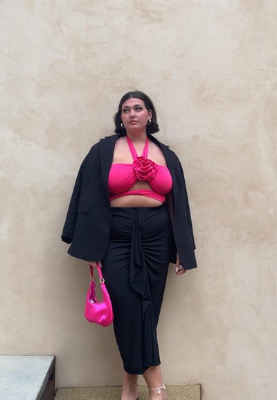 Mujer con cabello corto y oscuro que lleva un blazer negro y una falda, un top halter rosa brillante con detalle de rosa, sosteniendo un bolso rosa a juego, de pie contra una pared beige.