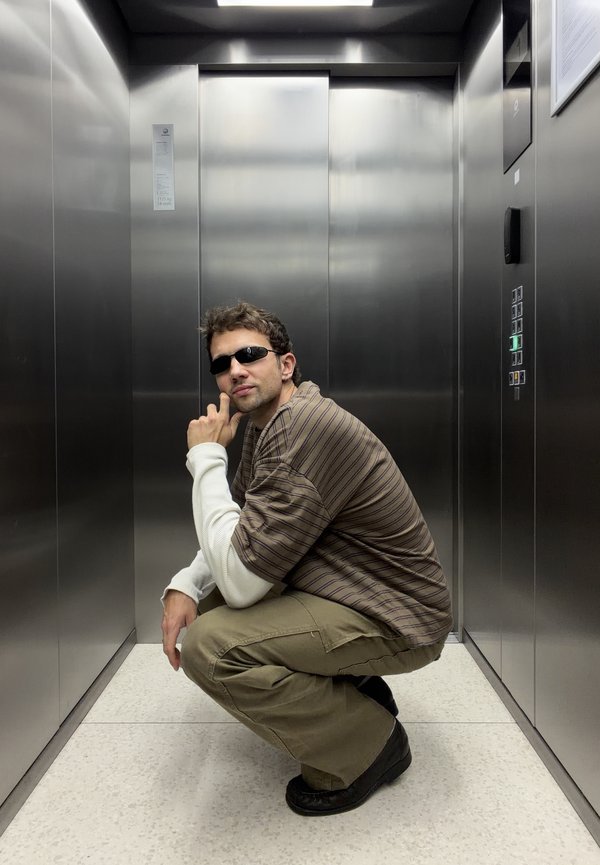 Hombre con gafas de sol agachado dentro de un ascensor moderno de acero inoxidable con puertas cerradas y botones de control iluminados.