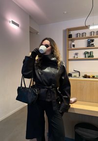 Femme portant une veste en cuir noire, des gants et des lunettes de soleil, buvant dans une tasse blanche dans une cuisine moderne avec des étagères et un comptoir en bois.