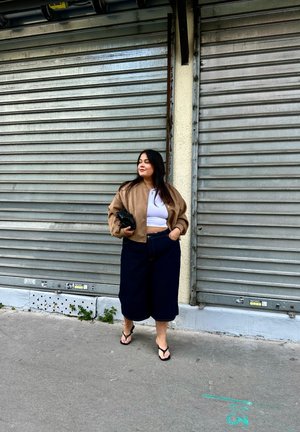 Tarnfarbene Bomberjacke, weißes Crop-Top, dunkelblaue Denim-Culottes und schwarze Sandalen. Steht vor einem welligen Metallhintergrund.