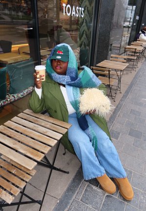 Persona con abrigo verde, bufanda azul, gorra verde y jeans sentada fuera de una cafetería sosteniendo una taza de café y una almohada beige esponjosa.