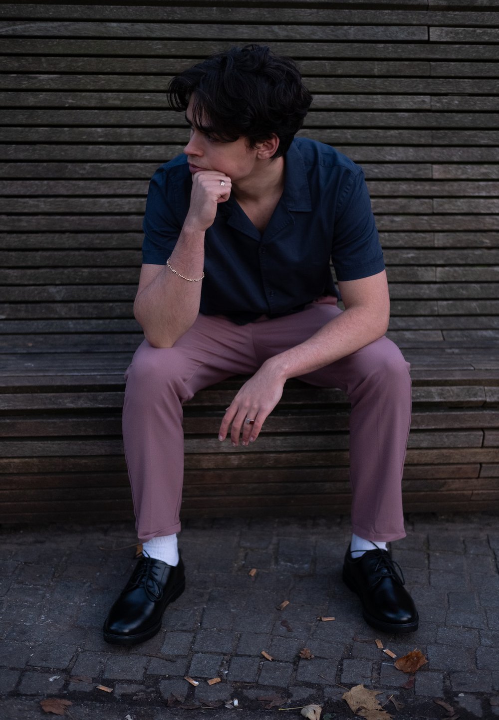 Man seated on a wooden bench, wearing a navy short-sleeved shirt and light pink trousers, paired with black shoes and white socks.