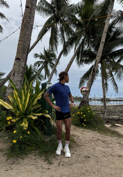Atleta con una camiseta azul de manga corta con cremallera y pantalones cortos negros, de pie cerca de palmeras, rodeado de plantas verdes y flores amarillas.